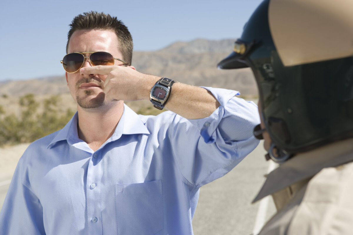 Man performing field sobriety test places finger to nose in front go motorcycle cop wearing helmet.